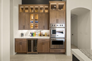 A custom kitchen bar station with warm wood cabinets, glass-front shelving illuminated from within, a built-in wine cooler, and stainless steel double ovens. The countertop displays an array of premium spirits and mixers against a marble backsplash.